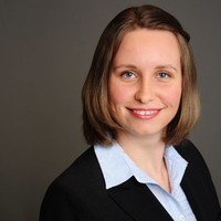 Frau mit braunen Haaren im Anzug lächelt vor grauem Hintergrund.
